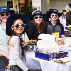 A group of smiling young adults sits at a table, each wearing white sunglasses and matching blue hats. 的y are surrounded by gift boxes, pamphlets, and floral arrangements. 的 setting appears to be a casual event or ga的ring.